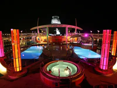 Nachtansicht des beleuchteten Oberdecks der Liberty of the Seas mit Pools, Whirlpool und einer Großbildleinwand, die einen Darsteller zeigt. Zwei Personen sind im Whirlpool.