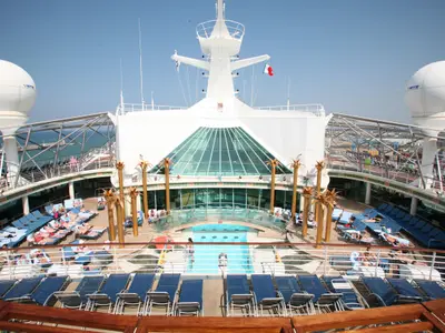 Das belebte Pooldeck der Liberty of the Seas mit blauen Sonnenliegen, Pools und einer großen Glasüberdachung. Radarkuppeln und ein Hafen sind im Hintergrund sichtbar.
