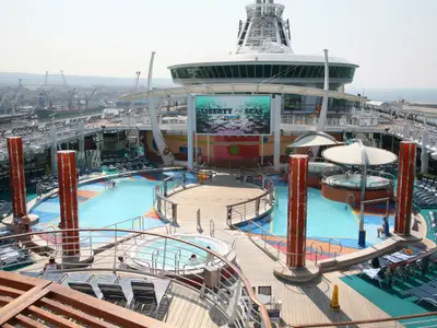 Belebtes Pooldeck der Liberty of the Seas mit Pools, Whirlpools und großem Bildschirm. Im Hintergrund ein Hafen mit Kränen unter hellem Tageslicht.