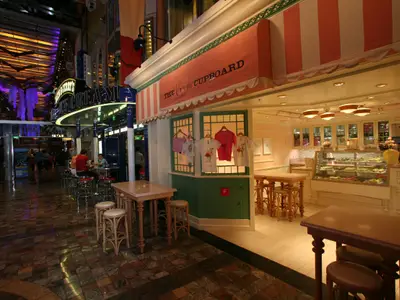 Die Royal Promenade der Liberty of the Seas. Links Ben & Jerry's, rechts The Cupcake Cupboard mit Auslagen. Gäste sitzen oder gehen durch die farbig beleuchtete Halle.