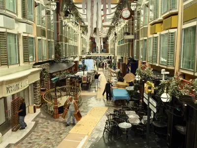Blick in die belebte Royal Promenade auf der Liberty of the Seas. Personal bereitet Geschäfte und Sitzbereiche entlang der mehrstöckigen Ladenzeile vor. Dekorationen hängen von der Decke.