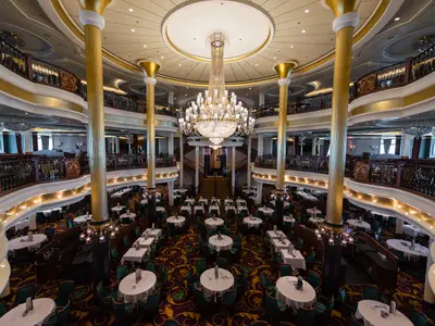 Das Hauptrestaurant der Liberty of the Seas, mehrstöckig mit großem Kronleuchter. Goldene Säulen und Balkone. Viele gedeckte Tische mit grünen Stühlen.