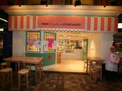 Eingang zu 'The Cupcake Cupboard' auf der Liberty of the Seas. Eine Mitarbeiterin im rosa Outfit steht rechts. Vor dem Laden stehen Holztische, drinnen Cupcakes in Auslagen.