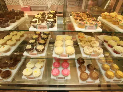 Die beleuchtete Gebäckvitrine auf der Liberty of the Seas, gefüllt mit appetitlichen Cupcakes. Reihen von Schokolade-, Vanille- und Karottenkuchen-Cupcakes auf weißen Platten präsentiert.