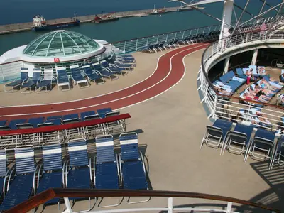 Blick von oben auf das Sonnendeck der Liberty of the Seas. Eine rote Laufstrecke und blaue Sonnenliegen, einige mit Passagieren, sind zu sehen. Im Hintergrund erstreckt sich ein Hafen mit Schiffen und einer Glaskuppel.