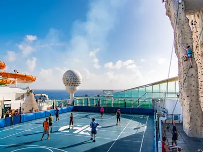 Deckansicht der Liberty of the Seas: Sportfeld, Kletterwand mit Kletterer, Wasserrutschen und Golfball-Skulptur unter blauem Himmel über dem Meer.