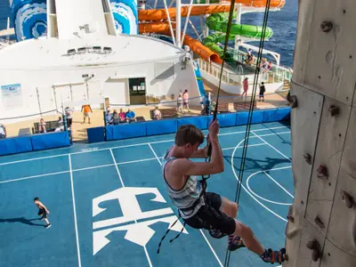 Ein junger Mann klettert an der Kletterwand auf dem Deck der Liberty of the Seas. Das Sportdeck mit Basketballfeld und weiteren Passagieren ist sichtbar, dahinter Wasserrutschen und das Meer.
