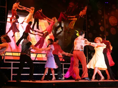 Eine Gruppe von Tänzern in farbenfrohen Kostümen performt eine choreografierte Show auf einer hell erleuchteten Bühne an Bord der Liberty of the Seas. Die Tänzer halten sich an Händen, mit Spiegelungen im Hintergrund, die die dynamische Szene erweitern.