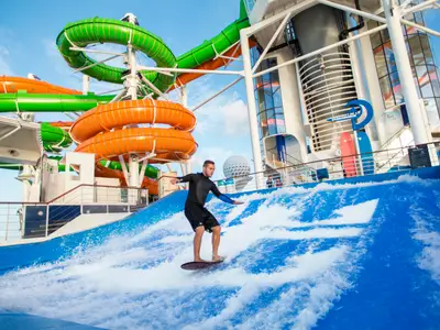 Mann surft auf dem FlowRider an Bord der Liberty of the Seas. Bunte Wasserrutschen und Aufbauten des Schiffes sind im Hintergrund unter blauem Himmel sichtbar.