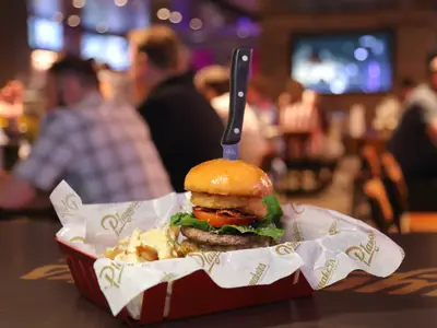 Nahaufnahme eines hohen Burgers mit Zwiebelringen und Speck, durchstochen von einem Steakmesser, dazu Pommes in einem roten Korb. Im Hintergrund verschwommen Gäste einer Sportsbar.