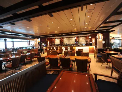 Geräumiger Restaurant- und Barbereich auf der Mariner of the Seas. Helle Holzmöbel, Korbstühle und eine lange Theke 'The Plaza Bar'. Personal im Service ist sichtbar.