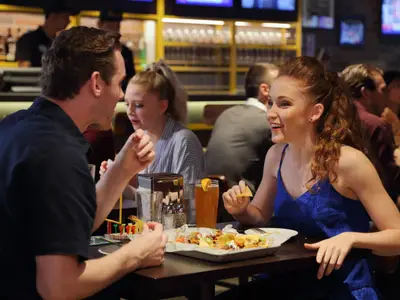 Ein Mann und eine Frau sitzen an einem Tisch in einer belebten Sportsbar. Die Frau in einem blauen Oberteil hält einen Nacho und lacht, während der Mann sie ansieht. Auf dem Tisch stehen Nachos, Getränke und ein kleines Tischspiel. Im Hintergrund sind weitere Gäste zu sehen.