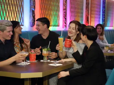 Eine Gruppe von fünf lachenden Freunden sitzt an einem Tisch in einer Tiki-Bar und trinkt bunte Cocktails. Die Innenwand ist mit Holzschnitzereien und farbigen Lichtern dekoriert.