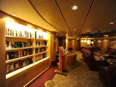 Eine gemütliche Bibliothek/Lounge auf der Mariner of the Seas mit beleuchteten Bücherregalen und roten Sofas. Ein Gast entspannt, ein Tresen ist sichtbar.
