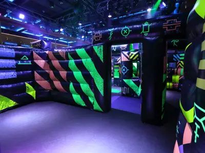 Blick in eine abgedunkelte Laser-Tag-Arena. Aufblasbare Wände und Hindernisse in Schwarz mit leuchtend grünen, orangefarbenen und blauen Mustern erzeugen ein futuristisches Ambiente. Ein Eingang führt tiefer in den Parcours.