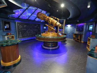 Raum auf der Mariner of the Seas mit großem goldenen Teleskop. Eine Sternendecke und Tafeln mit Formeln umrahmen das Szenario, akzentuiert von blauer Beleuchtung auf dem Boden.