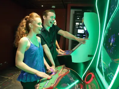 Ein junges Paar spielt begeistert an Arcade-Automaten in einem lebhaften Raum mit grünen LED-Akzenten. Der Mann zeigt auf den Bildschirm, die Frau bedient den Joystick.