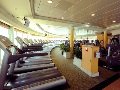 Fitnessstudio auf der Mariner of the Seas. Laufbänder an Fenstern, weitere Sportgeräte innen, Teppich, Säulen und Pflanzen schmücken den hellen Raum.