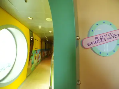Ein farbenfroher Flur auf der Mariner of the Seas mit einem runden Fenster, Kinder-Wandmalereien und einem Schild für 'Royal Babies and Tots'.