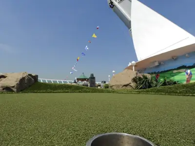Minigolf auf der Mariner of the Seas. Kunstrasen, Golf-Wandmalerei, Felsen, bunte Signalflaggen. Blauer Himmel, weiße Schiffsstruktur.