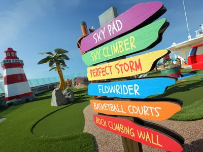 Outdoor-Freizeitdeck der Mariner of the Seas mit buntem Wegweiser zu Attraktionen wie FlowRider. Ein dekorativer Leuchtturm und Kunstrasen sind ebenfalls zu sehen.
