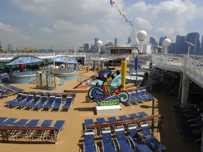 Blick auf das Pooldeck der Mariner of the Seas mit Sonnenliegen, Whirlpools und einem bunten Kunstwerk. Im Hintergrund eine Stadtsilhouette mit Kränen.