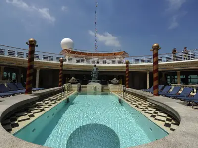 Sonniger Poolbereich der Mariner of the Seas: Länglicher Pool, Statue, Liegestühle. Obere Promenade mit zwei Personen. Radom, Flaggenmast, blauer Himmel.
