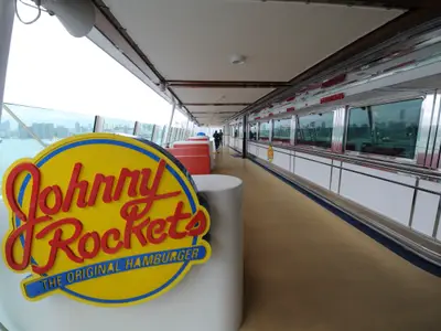Blick auf das Johnny Rockets Restaurant auf dem Promenadendeck der Mariner of the Seas. Gelbes Schild, Diner-Bereich mit Fenstern. Stadtkulisse im Hintergrund; Personen auf dem Deck.