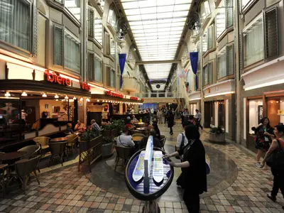 Die Royal Promenade an Bord der Mariner of the Seas, eine lebhafte Ladenstraße. Zahlreiche Passagiere flanieren oder sitzen in Cafés und Geschäften. Über den Etagen sind Kabinenfenster sichtbar, unter einem großen Glasdach.