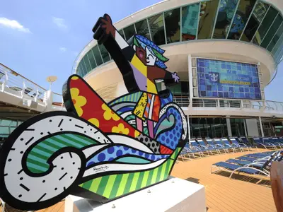 Bunte Skulptur auf dem Deck der Mariner of the Seas. Im Hintergrund sind das Royal Caribbean Logo auf einem Bildschirm und Liegestühle auf dem Holzdeck zu sehen.