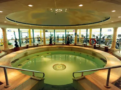 Blick auf den runden Innenpool und angrenzenden Fitnessbereich auf der Mariner of the Seas. Um den Pool verläuft eine Sitzbank. Im Hintergrund sind Fitnessgeräte und Fenster sichtbar.