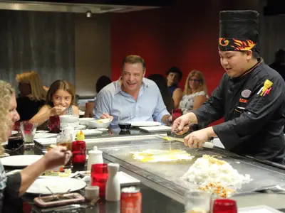 Ein asiatischer Koch in schwarzer Uniform und Stirnband bereitet an einer glänzenden Hibachi-Kochplatte Eier und Reis zu. Gäste, darunter eine lächelnde junge Frau und ein Mann, schauen begeistert zu und essen.