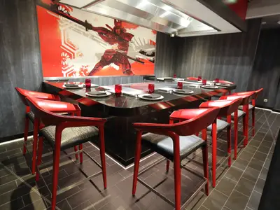 Innenansicht eines modernen Teppanyaki-Restaurants. Im Zentrum steht ein schwarzer Tisch mit Kochplatten und roten Barhockern. An der Wand prangt ein großes, rotes Samurai-Wandbild.