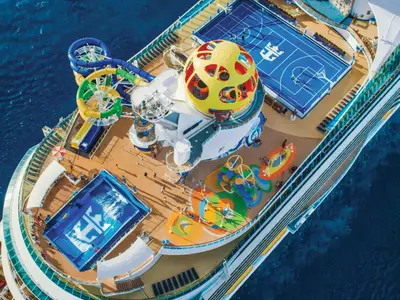Oberdeck der Mariner of the Seas von oben. Wasserrutschen, FlowRider, Basketballfeld, Spielbereich und gelbe Kugel zu sehen. Personen beleben das Deck, im blauen Meer.