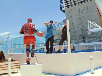 Das Sportdeck der Mariner of the Seas mit einer Basketballspieler-Statue. Zwei Männer montieren ein blaues Netz, daneben eine Kletterwand und ein Basketballkorb.