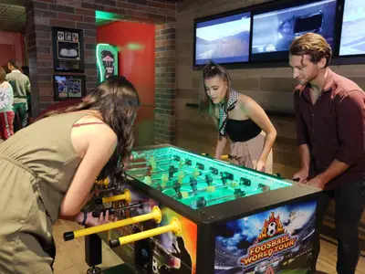 Drei junge Erwachsene spielen an einem modernen Foosball-Tisch mit grüner LED-Beleuchtung in einer lebhaften Sportbar. Im Hintergrund sind weitere Spielautomaten und Bildschirme zu sehen.