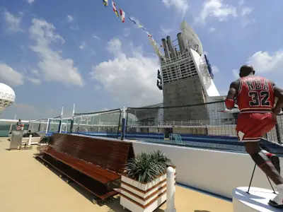 Sportdeck der Mariner of the Seas mit Statue eines Basketballspielers (Nr. 32) im Vordergrund, einem Sportfeld mit Netzen, einer Kletterwand und dem blauen Himmel.