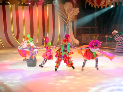 Eine Gruppe von fünf Eiskunstläufern in farbenfrohen Clownskostümen und einer Schmetterlingsfigur tritt auf einer beleuchteten Eisbühne auf. Ein weiterer Clown steht im Hintergrund auf der Bande.