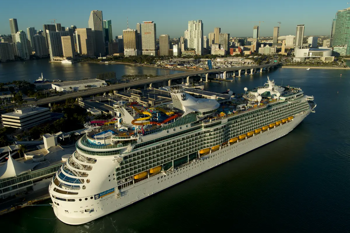 Luftbild zeigt die festgemachte Navigator of the Seas im Hafen von Miami, mit der imposanten Stadt-Skyline und einer Brücke im Hintergrund. Heller Sonnenschein.