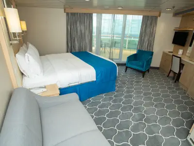 Innenansicht einer hellen Balkonkabine auf der Navigator of the Seas. Zu sehen sind ein Doppelbett, ein beiges Sofa, ein türkisblauer Sessel, ein Schreibtisch mit Stuhl und ein Balkon mit Meerblick.