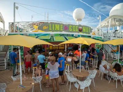 Gäste auf dem Deck der Navigator of the Seas tummeln sich an der Bar 'The Lime & Coconut'. Sie sitzen und stehen unter gelben Schirmen und genießen Getränke in entspannter Atmosphäre.