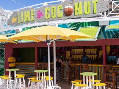 Außenbar 'The Lime & Coconut' auf der Navigator of the Seas. Gäste und Personal sind an der bunt gestreiften Theke zu sehen, umgeben von gelben Sonnenschirmen und Tischen.