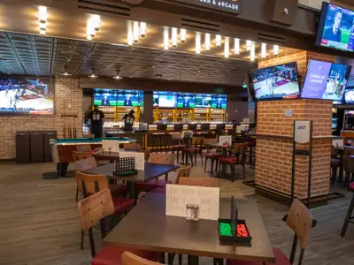 Eine moderne Sportbar auf der Navigator of the Seas. Zu sehen sind eine Bar mit Bildschirmen, Tische mit Spielen, ein Billardtisch und eine Ziegelsteinwand. Zwei Mitarbeiter stehen hinter der Bar.
