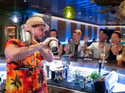 Ein Barkeeper schüttelt an der Bar der Navigator of the Seas Cocktails. Lachende Gäste schauen ihm begeistert zu. Flaschen, Gläser und Shaker sind auf der Theke zu sehen.
