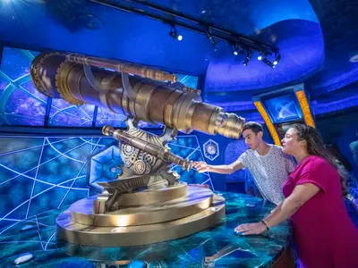 Ein junger Mann und eine Frau bedienen ein riesiges Steampunk-Teleskop in einem futuristischen, blau beleuchteten Raum. Bildschirme zeigen Weltall-Motive und geometrische Muster. Der Fokus liegt auf den Personen und dem Teleskop.