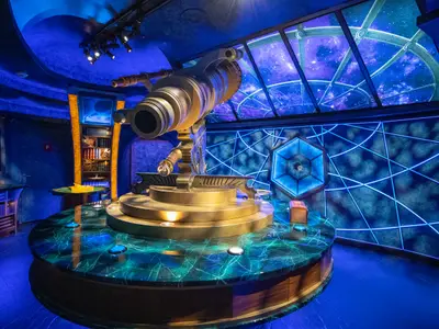 Innenansicht einer Sternwarte auf der Navigator of the Seas. Ein großes goldenes Teleskop steht zentral auf einem beleuchteten Tisch. Wände und Decke zeigen kosmische Projektionen in Blau- und Violetttönen.