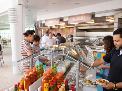 Mehrere Personen bedienen sich an einem hellen Buffet mit mexikanischem Essen. Eine Familie steht im Vordergrund, im Hintergrund ist das Küchenpersonal zu sehen. Viele Saucenflaschen stehen bereit.