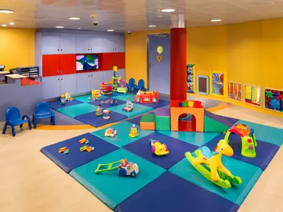 Ein farbenfroher Kinder-Spielbereich auf der Navigator of the Seas. Bunte Matten, diverse Spielzeuge, gelbe Wände, eine Schrankwand mit TV und kleine Stühle prägen das Bild.