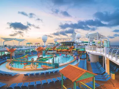 Blick auf das belebte Pooldeck der Navigator of the Seas bei Sonnenuntergang, mit mehreren Pools, einer Lazy River, vielen blauen Liegestühlen und bunten Bars.
