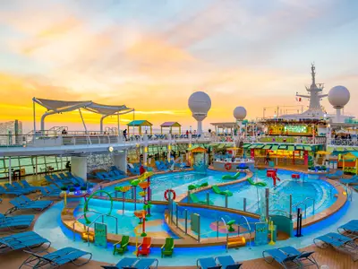 Sonnenuntergang auf dem Oberdeck der Navigator of the Seas. Die beleuchtete Poollandschaft mit Wasserspielen, bunten Bars und Liegestühlen ist gut sichtbar. Einige Personen sind an Bord.
